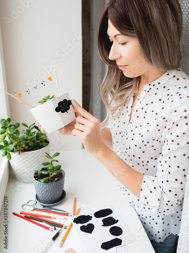 Smiling woman shows handmade decorations for Halloween. DIY flags and Boo! sticker on flowerpot with succulent plant. Socially-Distanced Halloween at home. photo