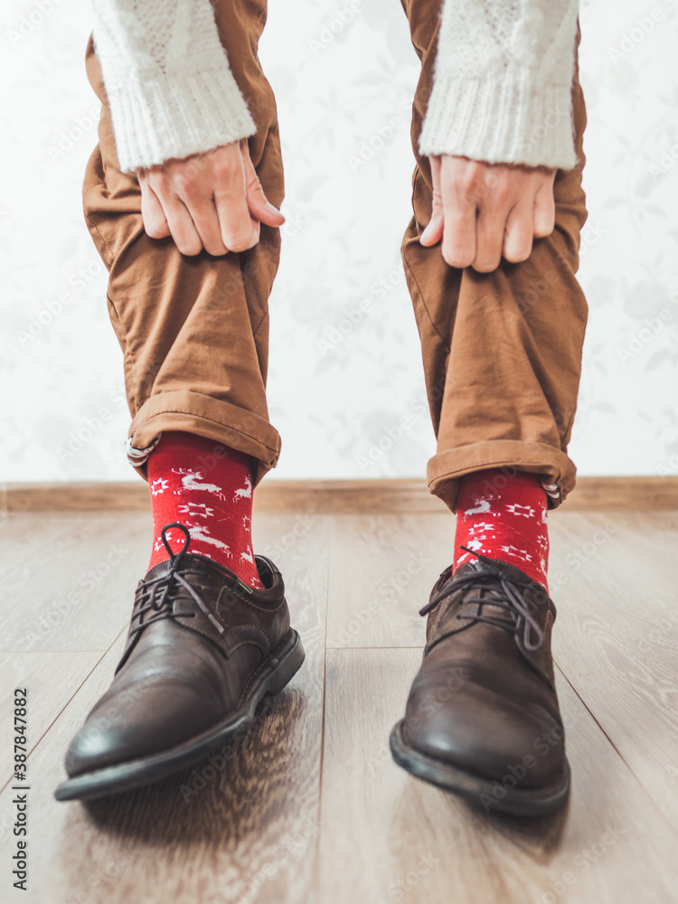 Young man pulls up leg of his chinos trousers to show bright red socks with  reindeers on them. Scandinavian pattern. Winter holiday spirit. Casual  outfit for New Year and Christmas celebration. Stock