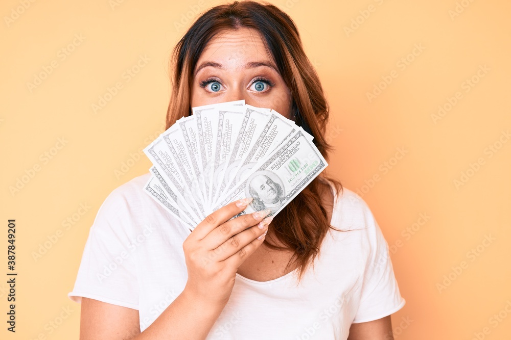 Young beautiful caucasian woman holding dollars covering mouth with hand, shocked and afraid for mistake. surprised expression Young beautiful caucasian woman holding dollars covering mouth with hand, shocked and afraid for mistake. surprised expression