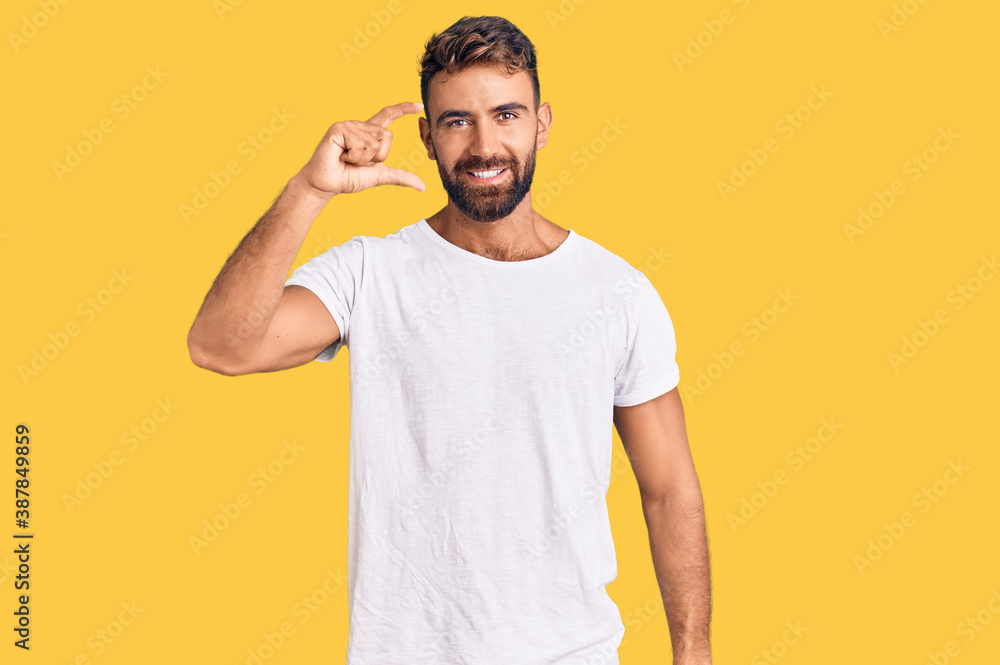 Young hispanic man wearing casual white tshirt smiling and confident gesturing with hand doing small size sign with fingers looking and the camera. measure concept. Young hispanic man wearing casual white tshirt smiling and confident gesturing with hand doing small size sign with fingers looking and the camera. measure concept.