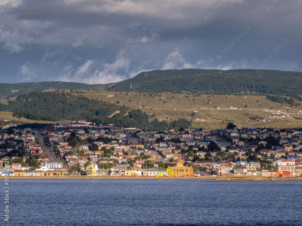 Punta Arenas, Chile - December 12, 2008: Sun shines on Cityscape of coastline centered on Hindu Temple and dark blue water, buildings with green hill on horizon under dark, storm, and rain cloudscape.