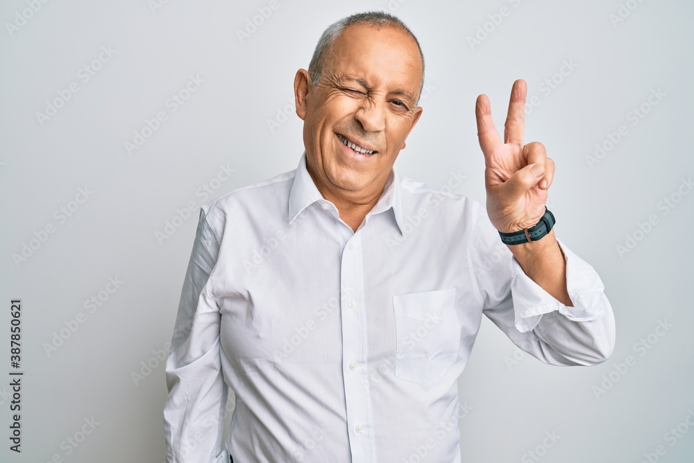 Handsome senior man wearing casual white shirt smiling with happy face winking at the camera doing victory sign with fingers. number two. Handsome senior man wearing casual white shirt smiling with happy face winking at the camera doing victory sign with fingers. number two.