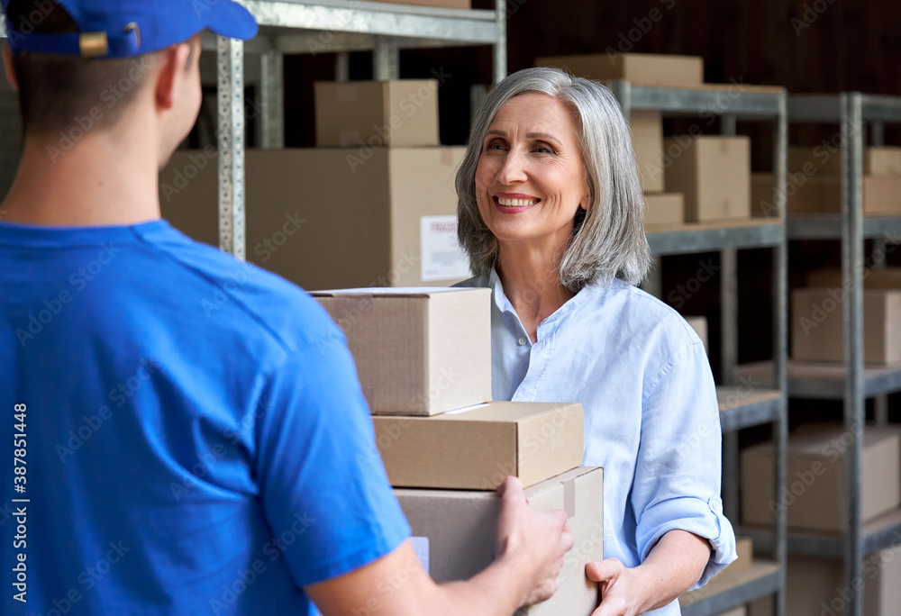 Obraz premium Happy mature female manager small business owner, seller, supervisor giving fast drop shipping safe delivery parcels packages boxes to male courier delivering ecommerce orders standing in warehouse.