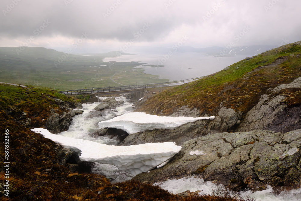 A bridge over a fast-flowing mountain stream, and snow on a Norwegian highland during a rainy and foggy summer day