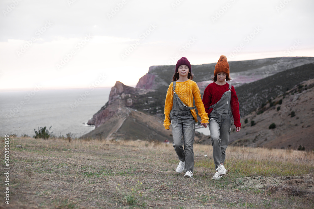 Two preteen girls traveler wearing denim overalls, yellow and red sweater and knitted hats sit on top of the mountain landscape and looking to the sea.