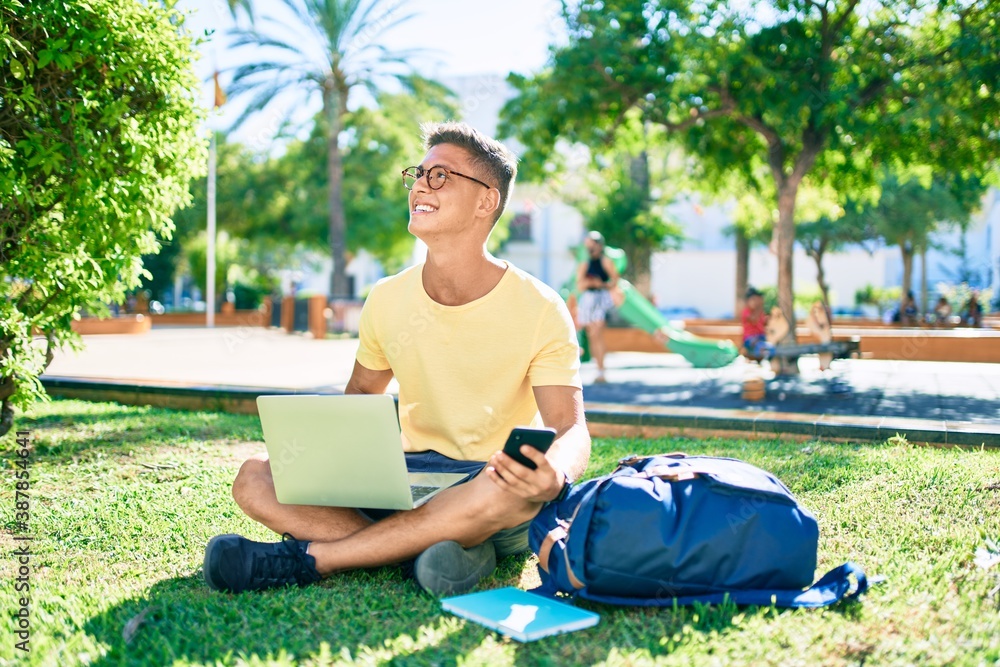 Obraz premium Young hispanic student smiling happy using laptop sitting on the grass at the university campus