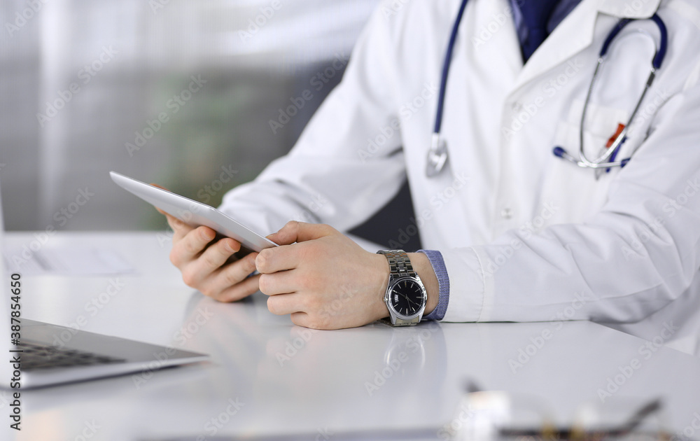 Unknown male doctor sitting and working with tablet computer in clinic at his working place, close-up. Young physician at work. Perfect medical service, medicine concept Unknown male doctor sitting and working with tablet computer in clinic at his working place, close-up. Young physician at work. Perfect medical service, medicine concept