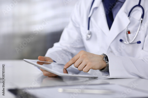 Unknown male doctor sitting and working with tablet computer in clinic at his working place, close-up. Young physician at work. Perfect medical service, medicine concept