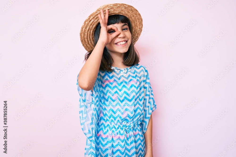 Young little girl with bang wearing summer dress and hat smiling happy doing ok sign with hand on eye looking through fingers Young little girl with bang wearing summer dress and hat smiling happy doing ok sign with hand on eye looking through fingers