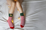 Female legs in Christmas socks with garland and festive lights in heart shape. Sexy woman on a bed, concept of romantic New Year night, elf costume