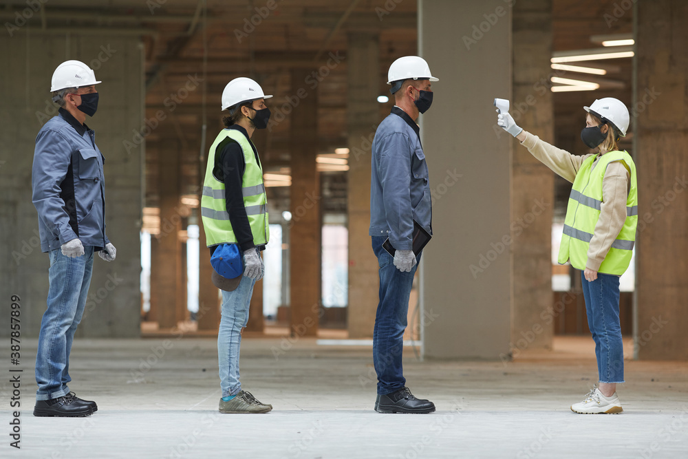 Side view full length of female supervisor measuring temperature of workers with contactless thermometer at construction site, corona virus safety measures Side view full length of female supervisor measuring temperature of workers with contactless thermometer at construction site, corona virus safety measures