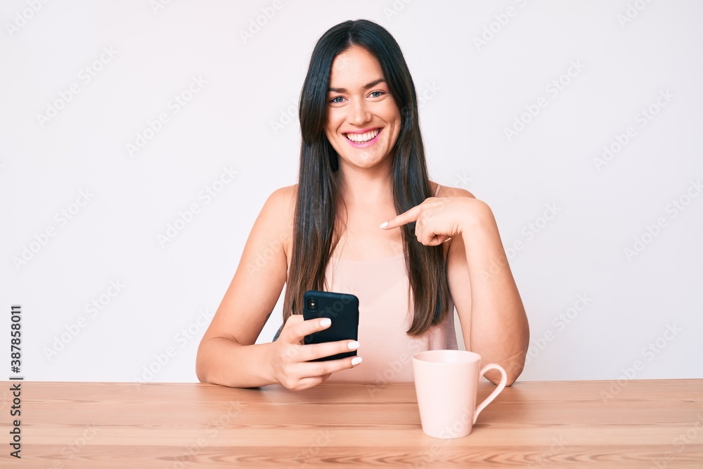 Young caucasian woman sitting at the desk using smartphone drinking coffee pointing finger to one self smiling happy and proud Young caucasian woman sitting at the desk using smartphone drinking coffee pointing finger to one self smiling happy and proud
