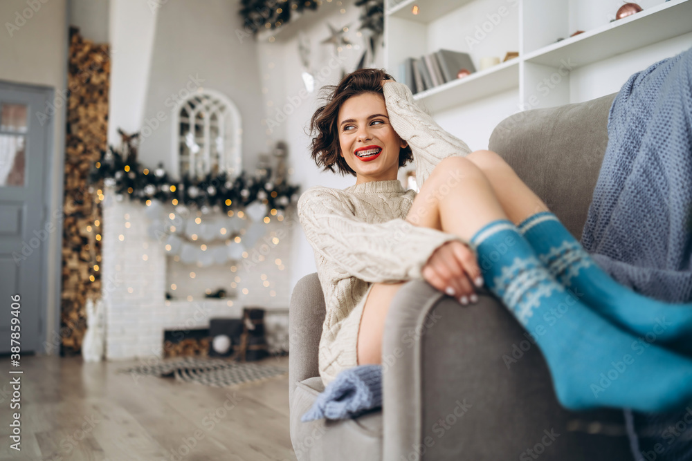 Pretty and happy brunette sitting in warm sweater on the chair with christmas lights behind. Pretty and happy brunette sitting in warm sweater on the chair with christmas lights behind.