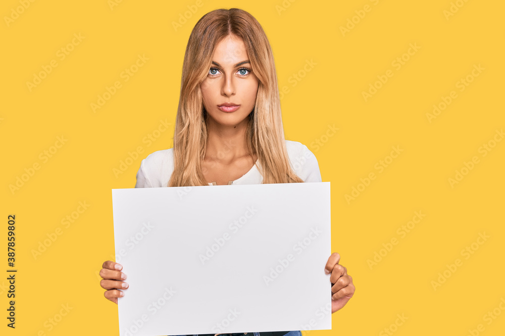 Naklejka premium Beautiful blonde young woman holding blank empty banner thinking attitude and sober expression looking self confident