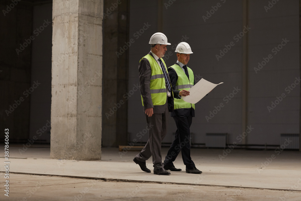 Full length portrait of two business people wearing hardhats and holding plans while walking at construction site indoors, copy space - obrazy, fototapety, plakaty 