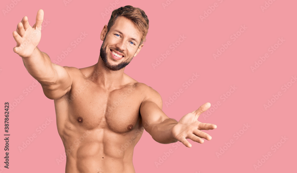 Fototapeta premium Young caucasian man standing shirtless looking at the camera smiling with open arms for hug. cheerful expression embracing happiness.