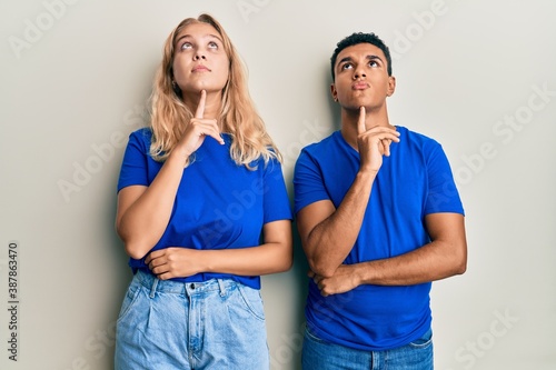 Young interracial couple wearing casual clothes thinking concentrated about doubt with finger on chin and looking up wondering