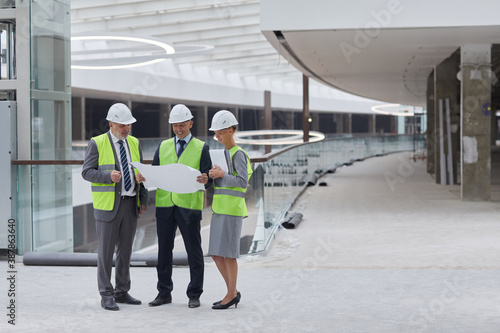 Full length portrait of three successful business people wearing hardhats and inspecting plans while standing at construction site indoors, copy space