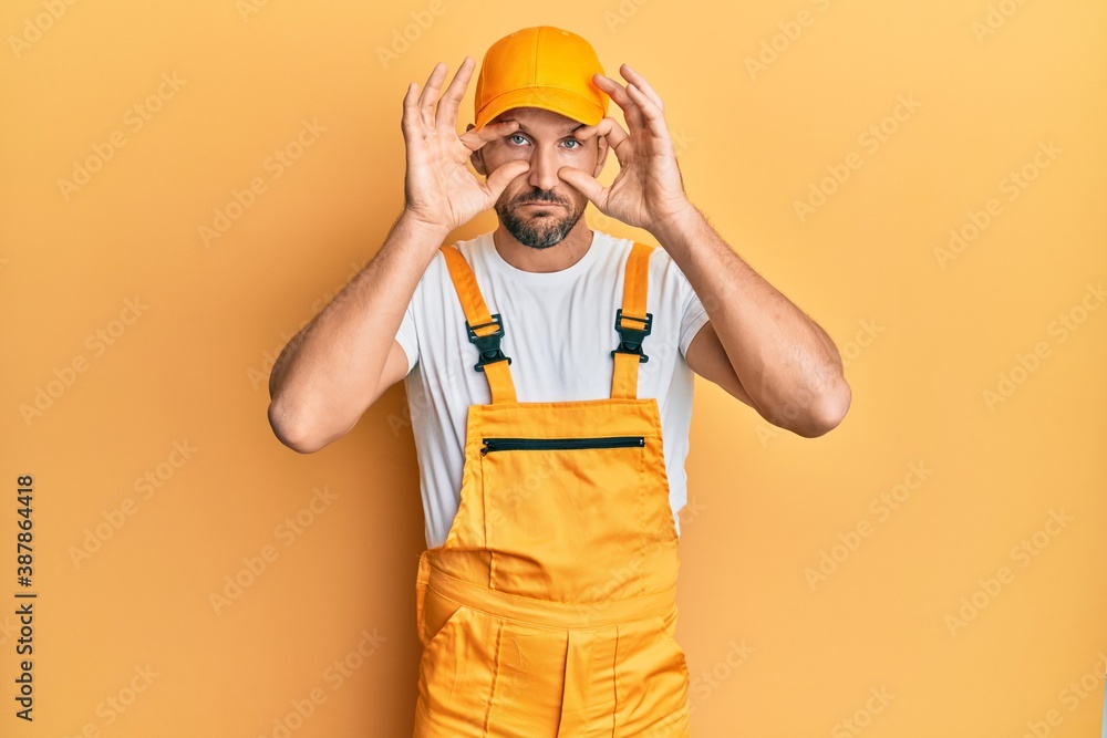 Young handsome man wearing handyman uniform over yellow background trying to open eyes with fingers, sleepy and tired for morning fatigue