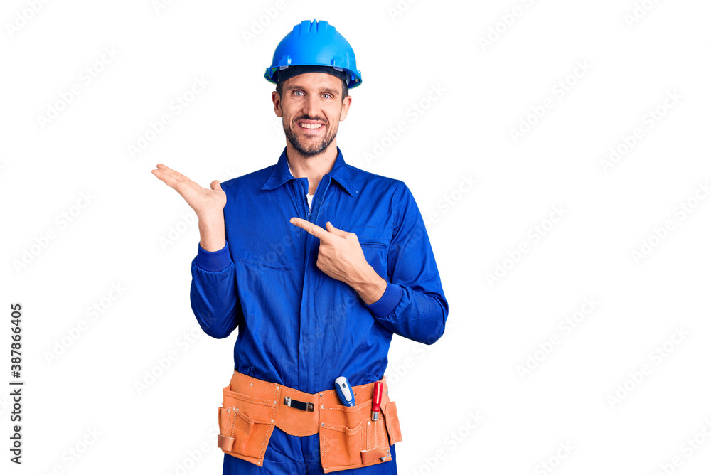 Young handsome man wearing worker uniform and hardhat amazed and smiling to the camera while presenting with hand and pointing with finger.