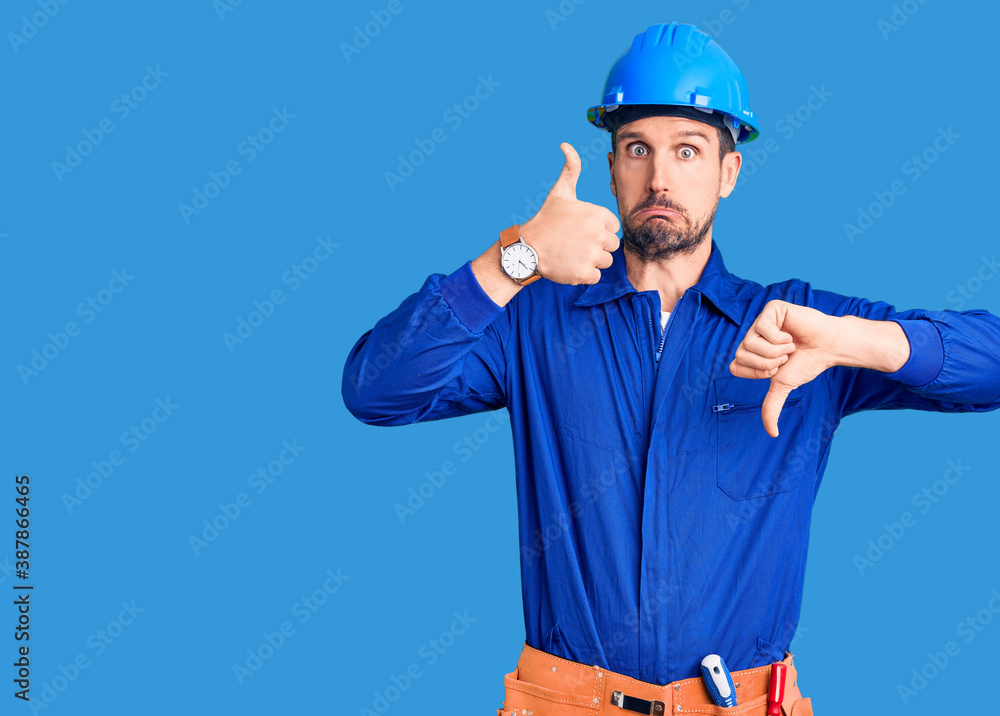 Young handsome man wearing worker uniform and hardhat pointing aside worried and nervous with both hands, concerned and surprised expression