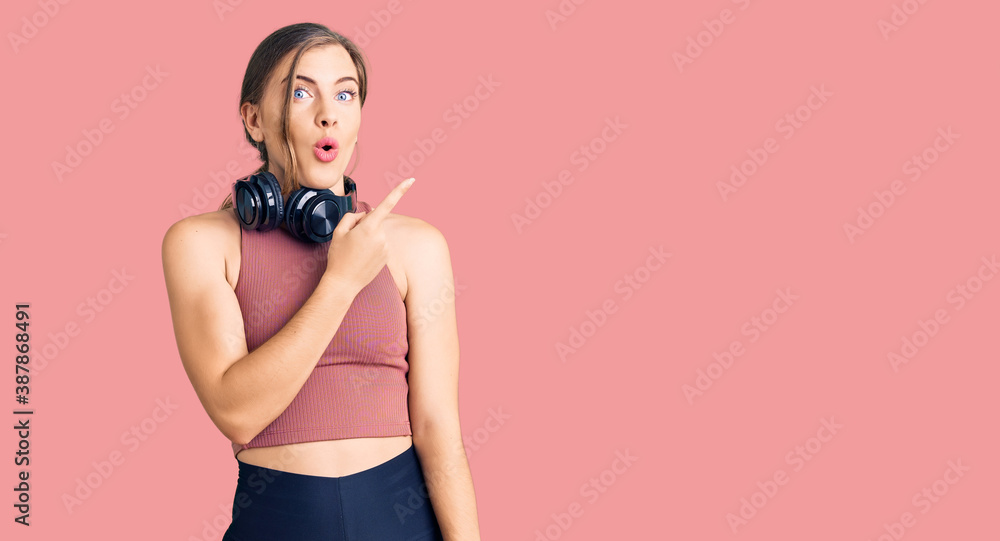 Obraz premium Beautiful caucasian young woman wearing gym clothes and using headphones surprised pointing with finger to the side, open mouth amazed expression.