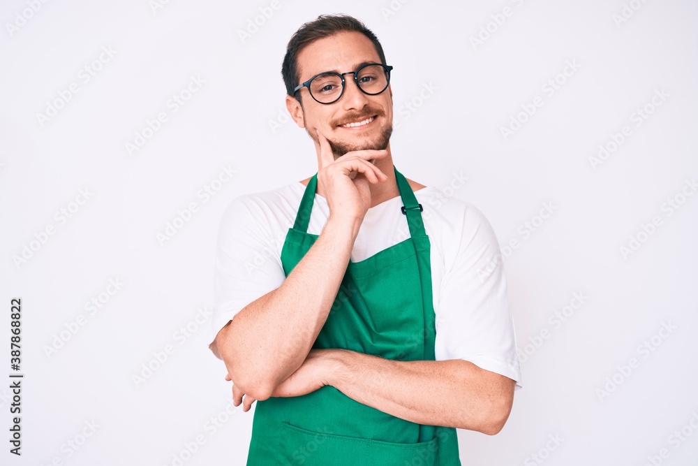 Young handsome man wearing apron smiling looking confident at the camera with crossed arms and hand on chin. thinking positive. Young handsome man wearing apron smiling looking confident at the camera with crossed arms and hand on chin. thinking positive.