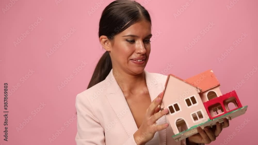 happy young businesswoman in pink suit holding and recommending house model, nodding and approving on pink background happy young businesswoman in pink suit holding and recommending house model, nodding and approving on pink background