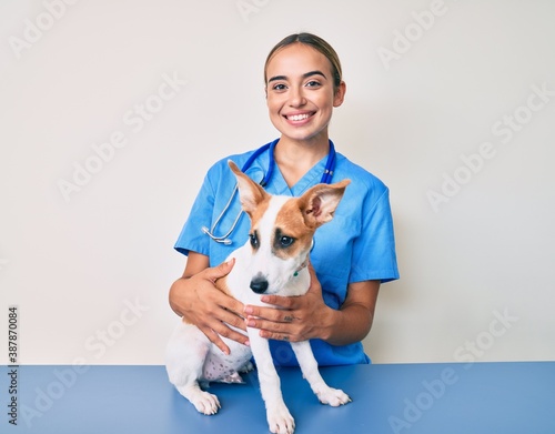 Young beautiful blonde veterinarian woman checking dog health looking positive and happy standing and smiling with a confident smile showing teeth photo
