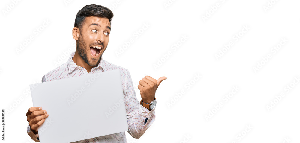 Handsome hispanic man holding blank empty banner pointing thumb up to the side smiling happy with open mouth Handsome hispanic man holding blank empty banner pointing thumb up to the side smiling happy with open mouth