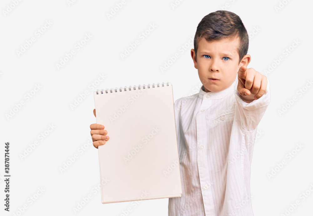 Cute blond kid holding notebook pointing with finger to the camera and to you, confident gesture looking serious Cute blond kid holding notebook pointing with finger to the camera and to you, confident gesture looking serious