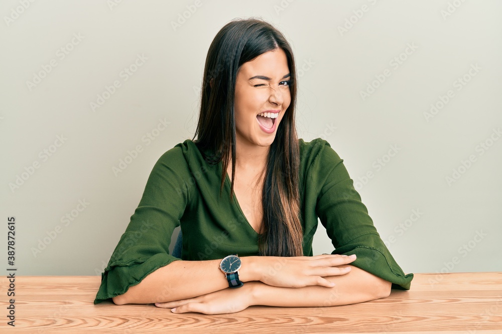 Young brunette woman wearing casual clothes sitting on the table winking looking at the camera with sexy expression, cheerful and happy face. Young brunette woman wearing casual clothes sitting on the table winking looking at the camera with sexy expression, cheerful and happy face.