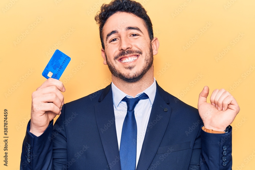 Young hispanic man wearing suit holding credit card screaming proud, celebrating victory and success very excited with raised arm Young hispanic man wearing suit holding credit card screaming proud, celebrating victory and success very excited with raised arm
