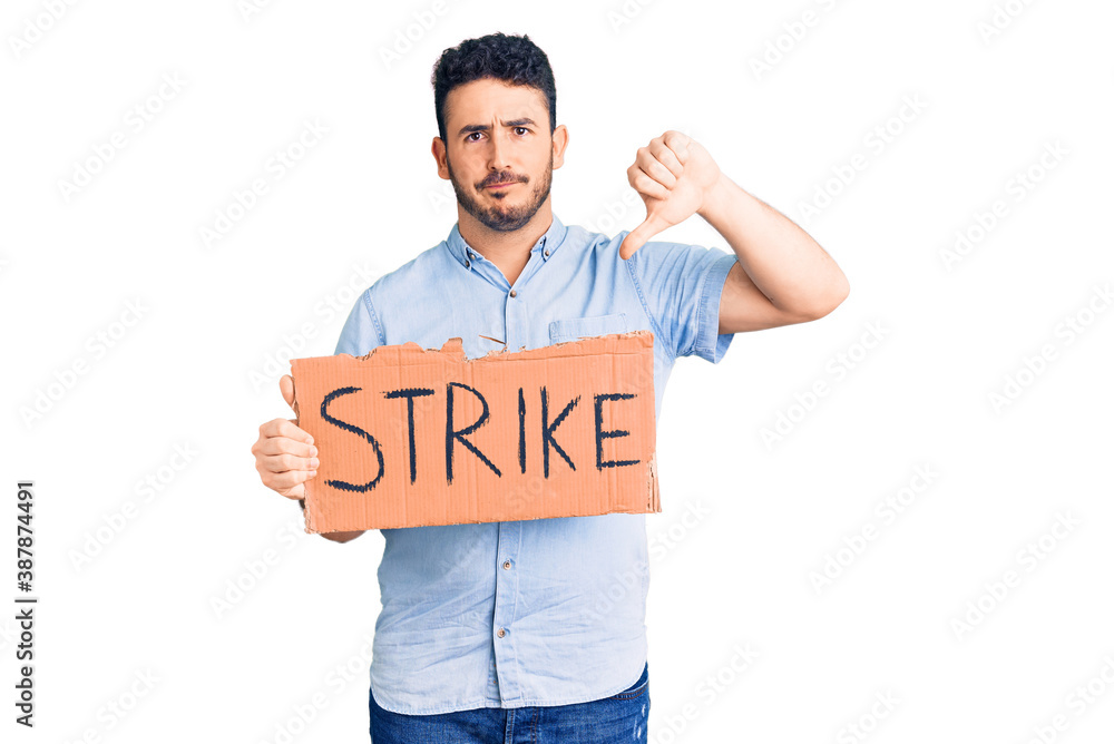 Young hispanic man holding strike banner cardboard with angry face, negative sign showing dislike with thumbs down, rejection concept Young hispanic man holding strike banner cardboard with angry face, negative sign showing dislike with thumbs down, rejection concept