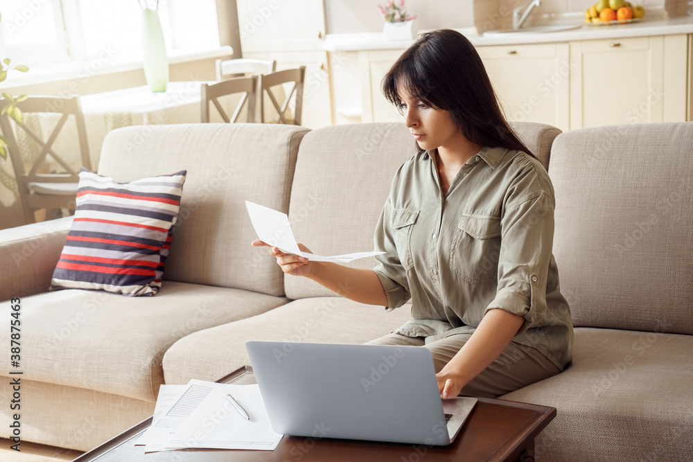 Young indian woman holding letter using laptop computer application paying bill online on website, managing account finances, reviewing paycheck banking loan debt payment sitting on couch at home. Young indian woman holding letter using laptop computer application paying bill online on website, managing account finances, reviewing paycheck banking loan debt payment sitting on couch at home.