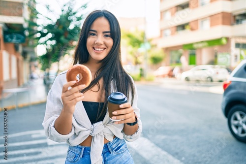 Young hispanic girl having breakfast eating donut and drinking take away coffee at the city.