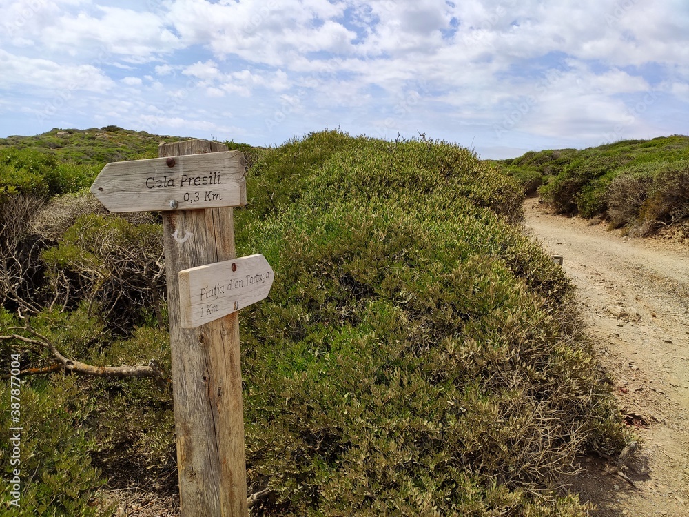 Wooden trail signpost arrow pointing to famous Cala Presili Beach on the north coast of Menorca Island with wild vegatation in background, near Far de Favaritx (Cap Favaritx), Balearic Islands, Spain