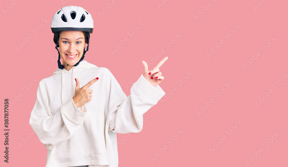 Beautiful brunette young woman wearing bike helmet and sporty clothes smiling and looking at the camera pointing with two hands and fingers to the side. Beautiful brunette young woman wearing bike helmet and sporty clothes smiling and looking at the camera pointing with two hands and fingers to the side.