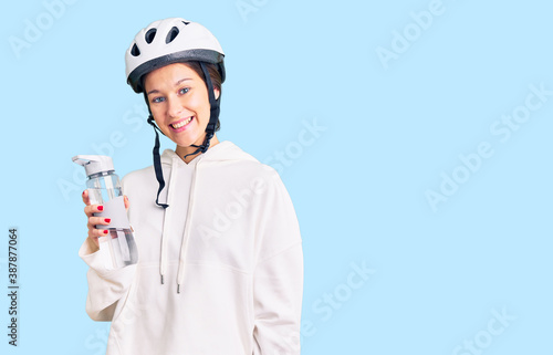 Beautiful brunette young woman wearing bike helmet and holding water bottle looking positive and happy standing and smiling with a confident smile showing teeth