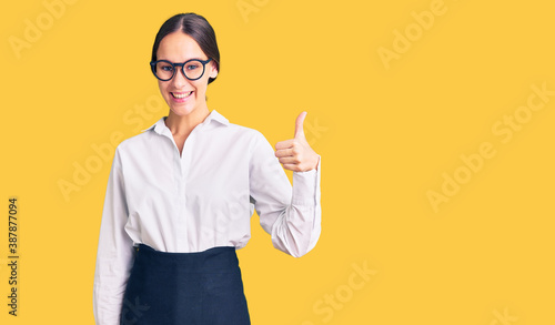 Beautiful brunette young woman wearing professional waitress apron doing happy thumbs up gesture with hand. approving expression looking at the camera showing success.