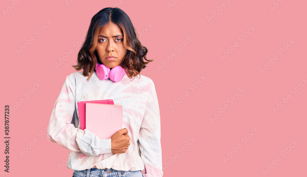 Young beautiful mixed race woman holding books wearing headphones thinking attitude and sober expression looking self confident Young beautiful mixed race woman holding books wearing headphones thinking attitude and sober expression looking self confident