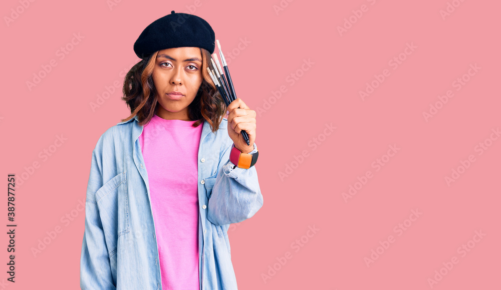 Young beautiful mixed race woman holding paintbrushes thinking attitude and sober expression looking self confident Young beautiful mixed race woman holding paintbrushes thinking attitude and sober expression looking self confident