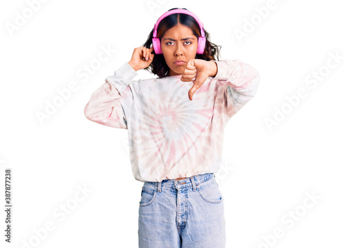 Young beautiful mixed race woman listening to music using headphones with angry face, negative sign showing dislike with thumbs down, rejection concept © Krakenimages.com