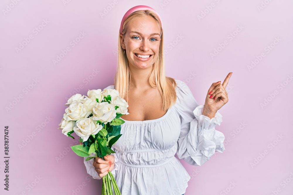 Young blonde girl holding flowers smiling happy pointing with hand and finger to the side
