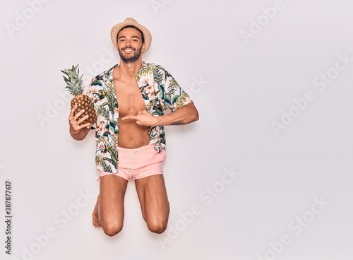 Young handsome hispanic man on vacation wearing swimwear, floral shirt and hat smiling happy. Jumping with smile on face holding pineapple over isolated white background
