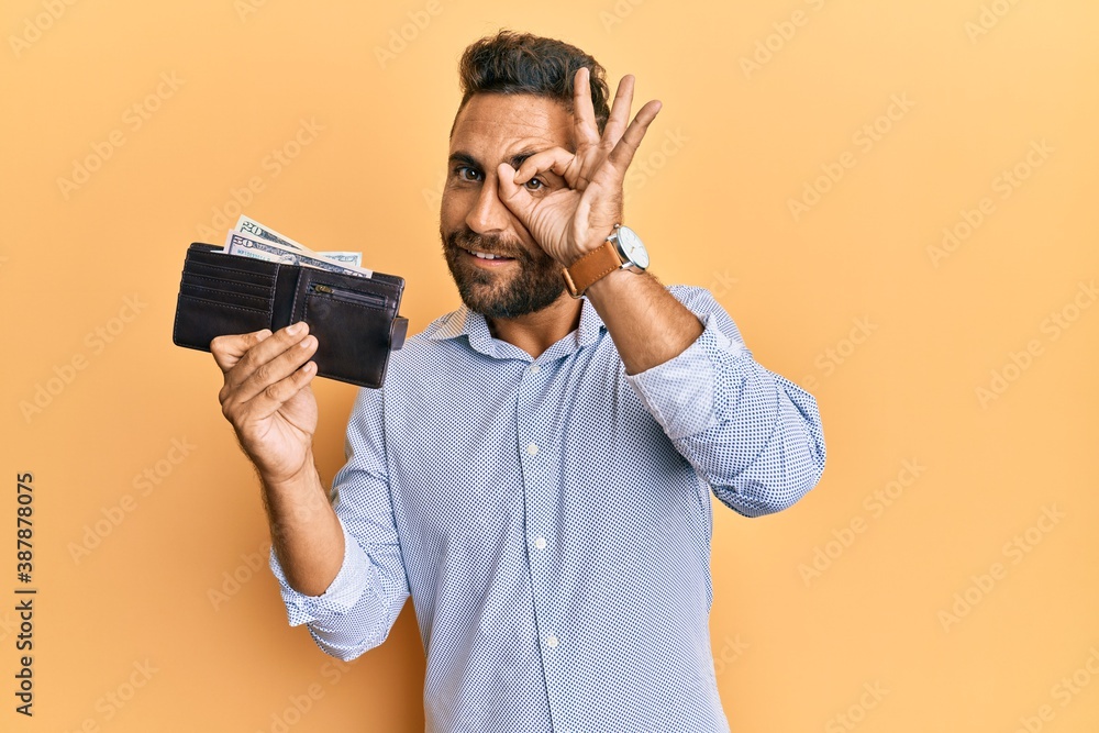 Handsome man with beard holding wallet with dollars smiling happy doing ok sign with hand on eye looking through fingers Handsome man with beard holding wallet with dollars smiling happy doing ok sign with hand on eye looking through fingers