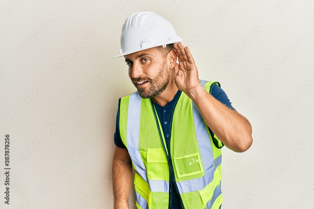 Handsome man with beard wearing safety helmet and reflective jacket smiling with hand over ear listening and hearing to rumor or gossip. deafness concept. Handsome man with beard wearing safety helmet and reflective jacket smiling with hand over ear listening and hearing to rumor or gossip. deafness concept.