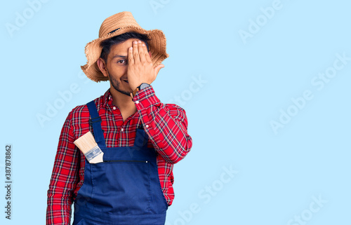 Handsome latin american young man weaing handyman uniform covering one eye with hand, confident smile on face and surprise emotion. photo