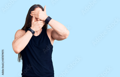 Young adult man with long hair wearing goth style with black clothes covering eyes and mouth with hands, surprised and shocked. hiding emotion
