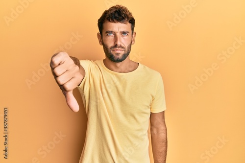 Handsome man with beard wearing casual yellow tshirt over yellow background looking unhappy and angry showing rejection and negative with thumbs down gesture. bad expression.